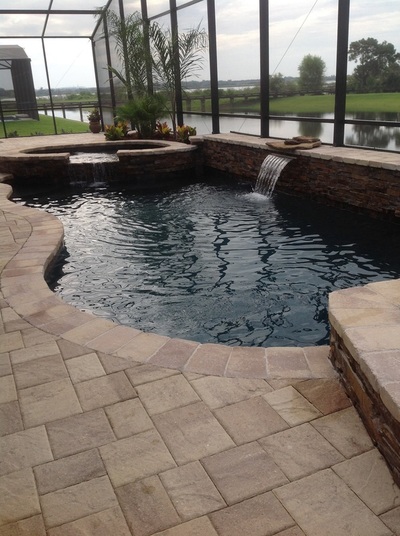 A freeform pool with a raised spa and waterfall spilling into dark water under a screened enclosure overlooking a lake.