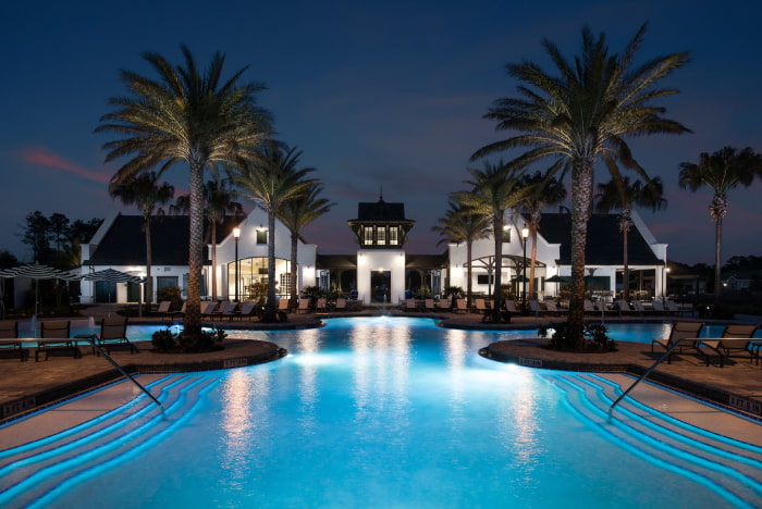 A large resort-style pool glowing blue at night in front of an illuminated clubhouse framed by palm trees.