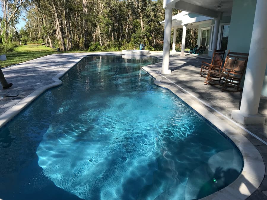 An L-shaped concrete pool with a raised spa behind a covered patio with rocking chairs.