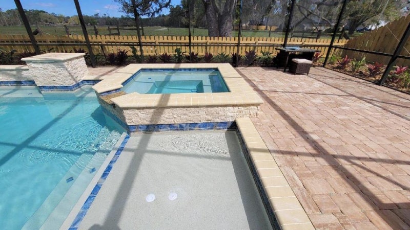 A raised spa with blue tile trim sits beside a pool's tanning shelf under a screened patio enclosure.