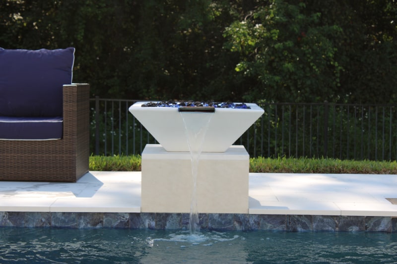 A raised white water bowl feature pours a stream into the pool beside a wicker lounge chair.