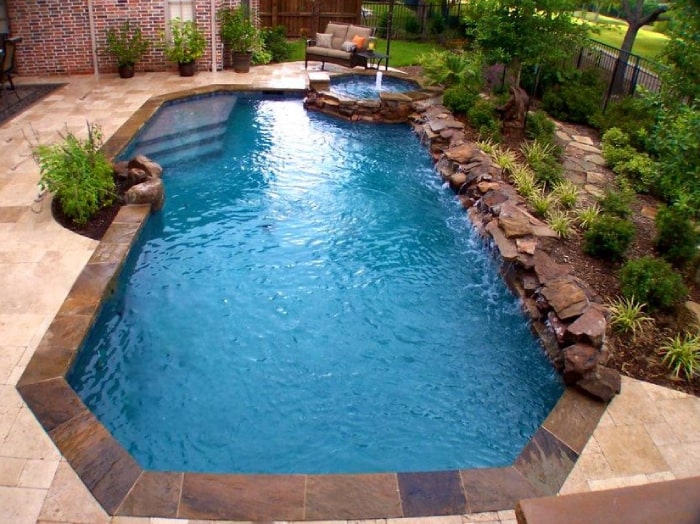 A small freeform backyard pool with a raised rock spa spilling into it, surrounded by travertine decking and landscaping.