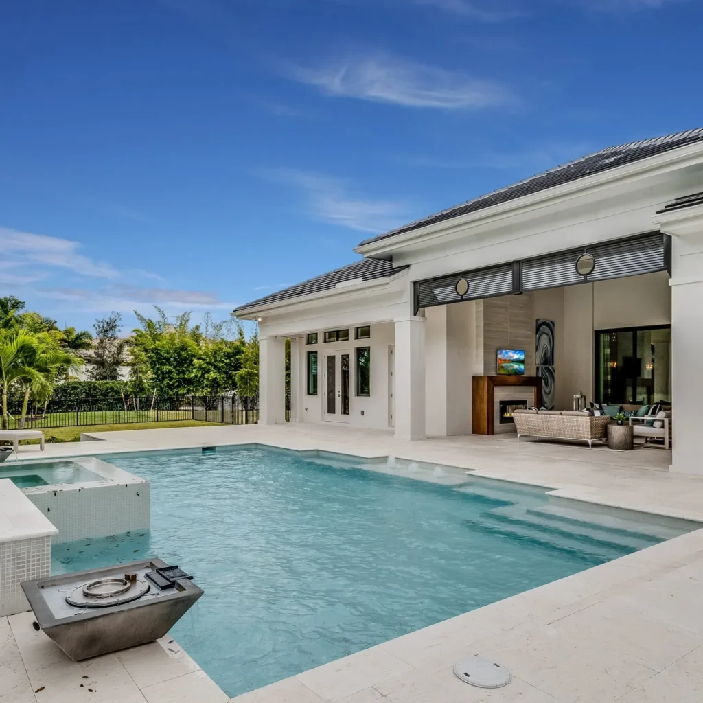 A rectangular pool with a raised tiled spa sits on a stone patio beside an open-air outdoor living room with fireplace.