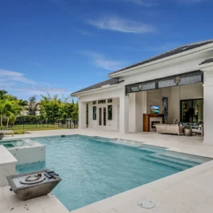 A rectangular pool with a raised tiled spa sits on a stone patio beside an open-air outdoor living room with fireplace.