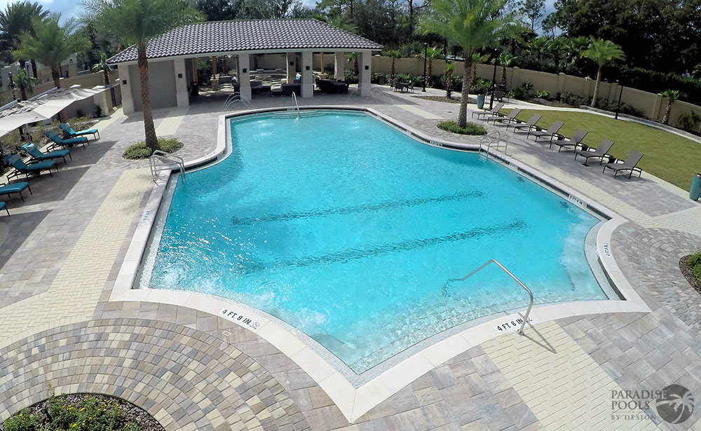 Aerial view of a large geometric community pool surrounded by paver decking, loungers, and a cabana.