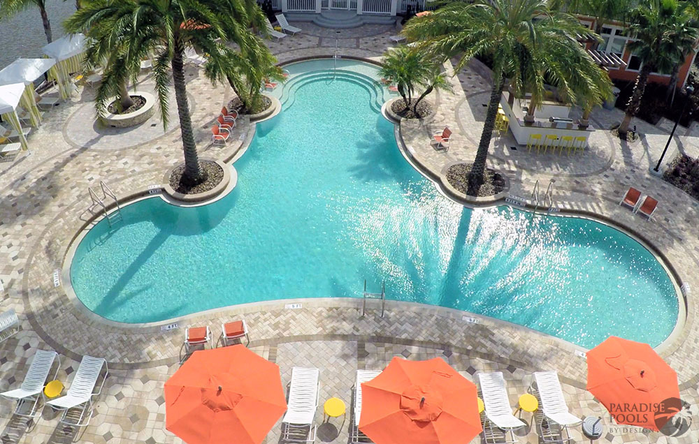 Aerial view of a large freeform resort pool surrounded by palm trees, orange umbrellas, and lounge chairs.