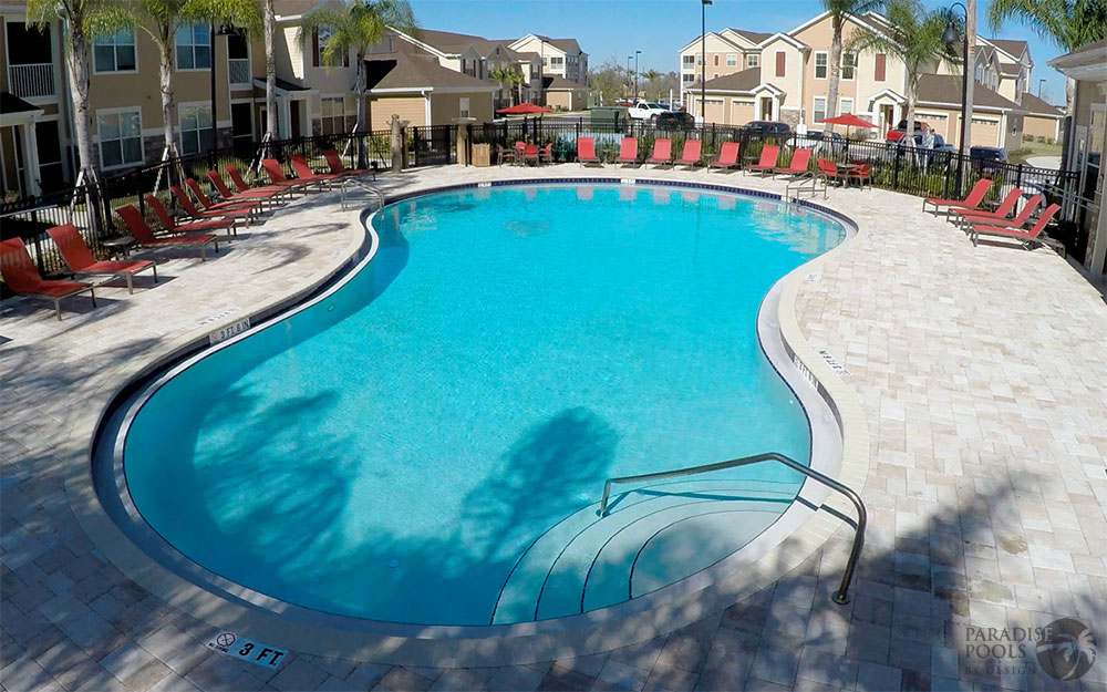 A freeform community pool surrounded by travertine decking and red lounge chairs, with apartment buildings behind.