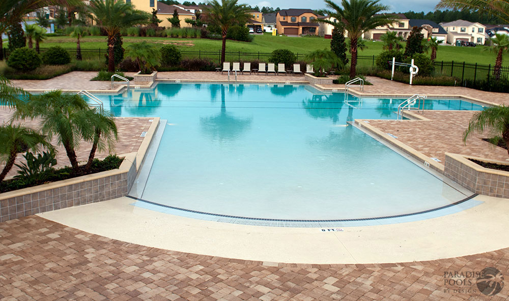 A large community pool with a zero-depth beach entry surrounded by paver decking and neighborhood homes.