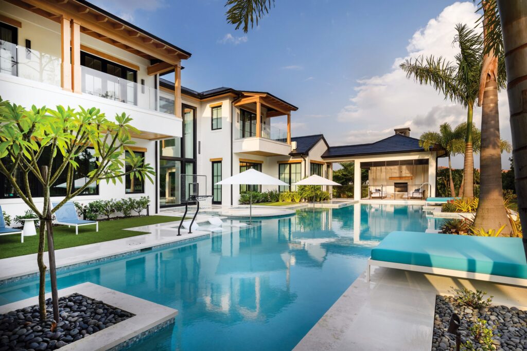 A long modern lap pool runs alongside a luxury two-story home with palm trees, umbrellas, and a poolside basketball hoop.