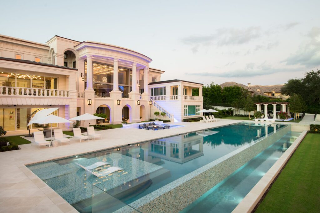 A long lap pool with a glass-walled elevated spa runs alongside a grand mansion at dusk.