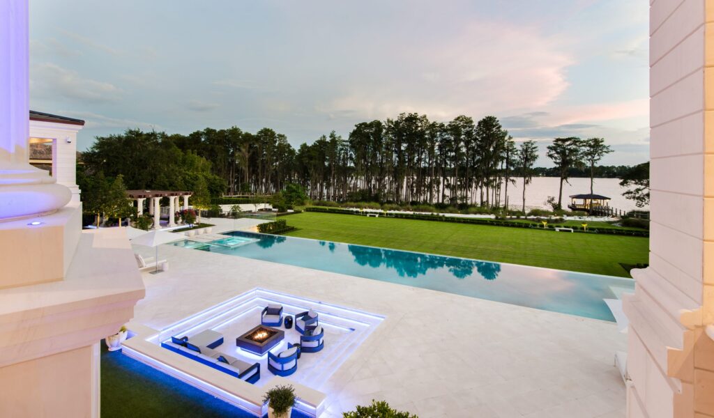 An infinity-edge lap pool overlooks a lake at dusk with a raised spa and a sunken fire pit lounge on the terrace.