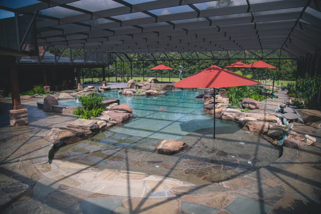 A large freeform pool with natural rock accents and a beach entry under a screened enclosure, shaded by red umbrellas.