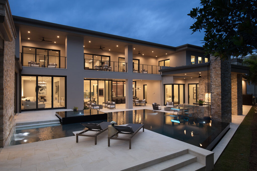 A modern two-story home at dusk with a sleek dark infinity-edge pool and raised knife-edge water feature on a stone patio.