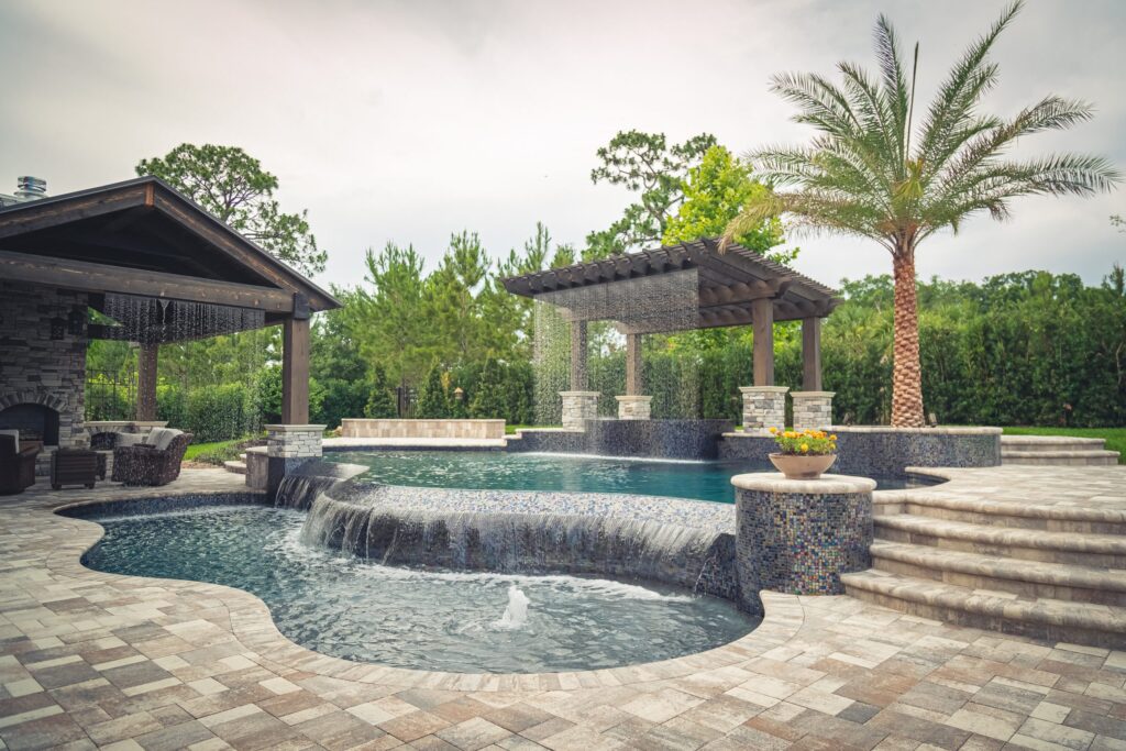 A freeform pool with a raised tiled spa, rain curtain feature, and pergola beside a paver patio.