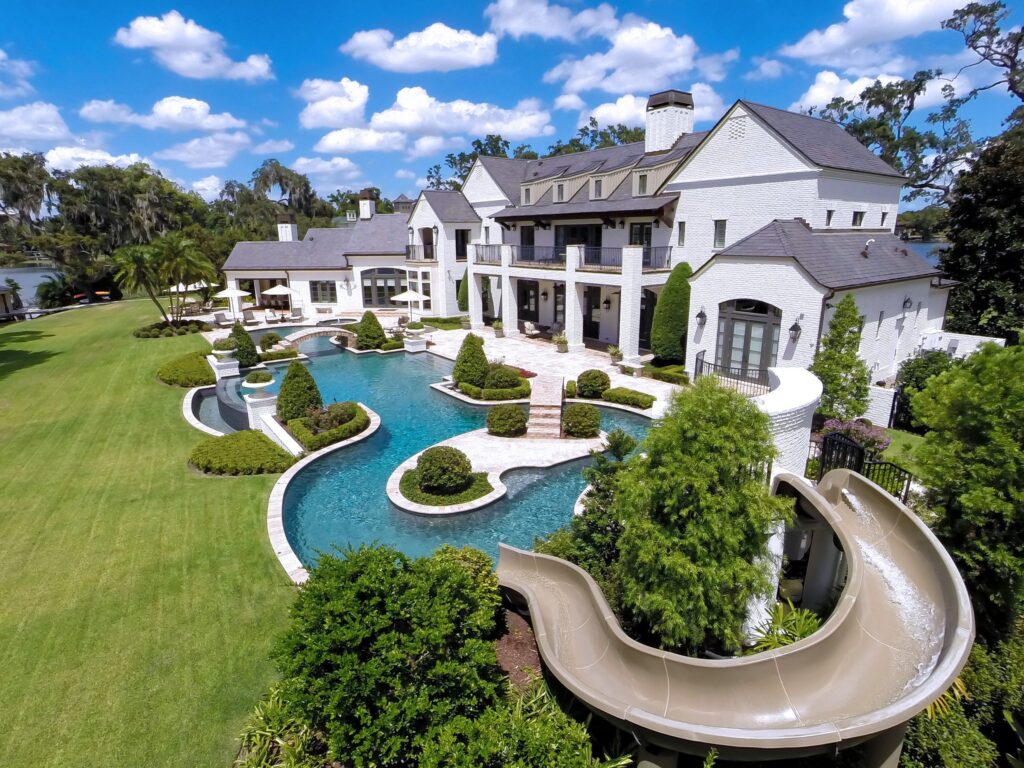 Aerial view of a sprawling freeform pool with islands and a curving water slide behind a large white estate home on the water.