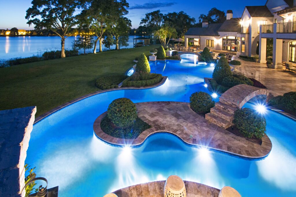 An elaborate freeform pool with stone bridge and planter islands glows blue at dusk beside a lake.