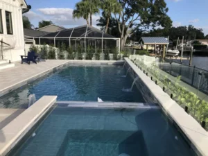 A rectangular infinity-edge style pool with a raised spa and water scuppers overlooking a canal with boats docked behind glass railing.