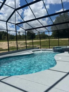 A freeform pool with a raised stone spa sits under a screened enclosure overlooking an open field.