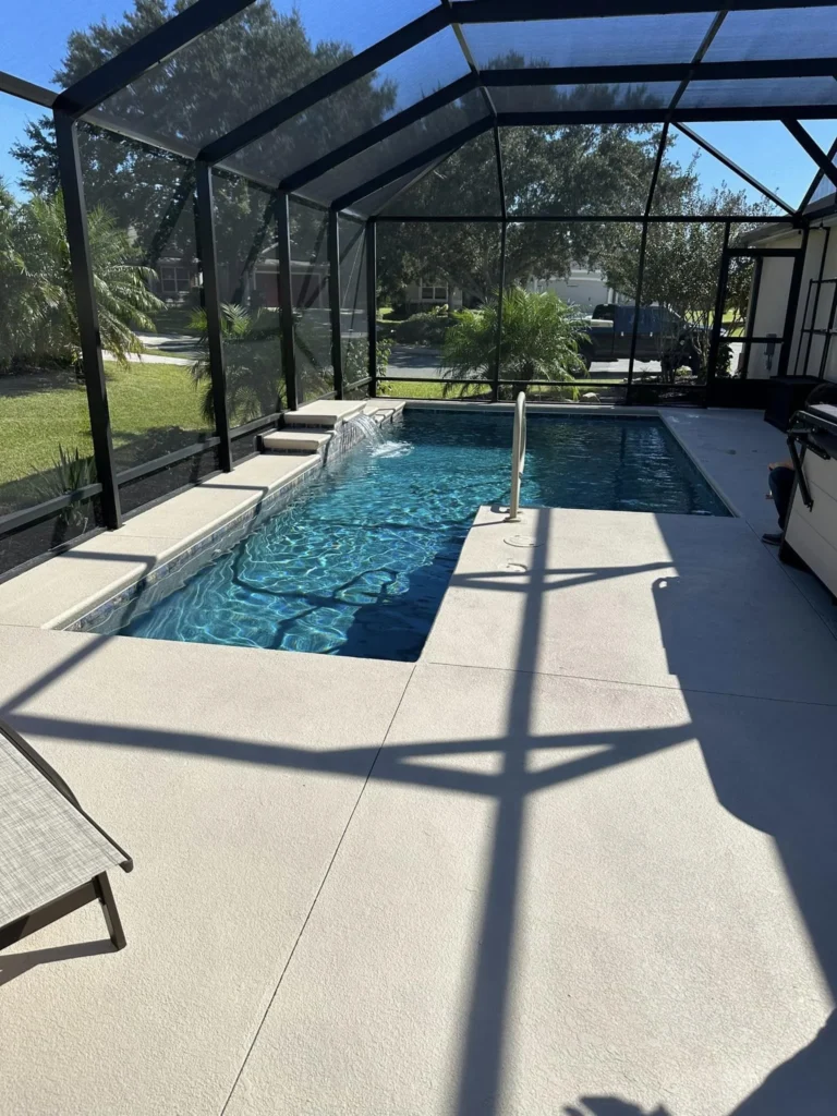 A screened rectangular backyard pool with a small water feature and step, seen through the pool cage on a sunny day.