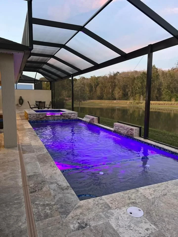 A rectangular pool with purple LED lighting and a raised spa with sheer descent waterfalls, enclosed in a screened lanai overlooking a pond at dusk.