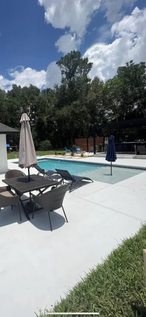 A small rectangular backyard pool with a concrete patio, lounge chairs, and a wooden pergola nearby under a sunny sky.
