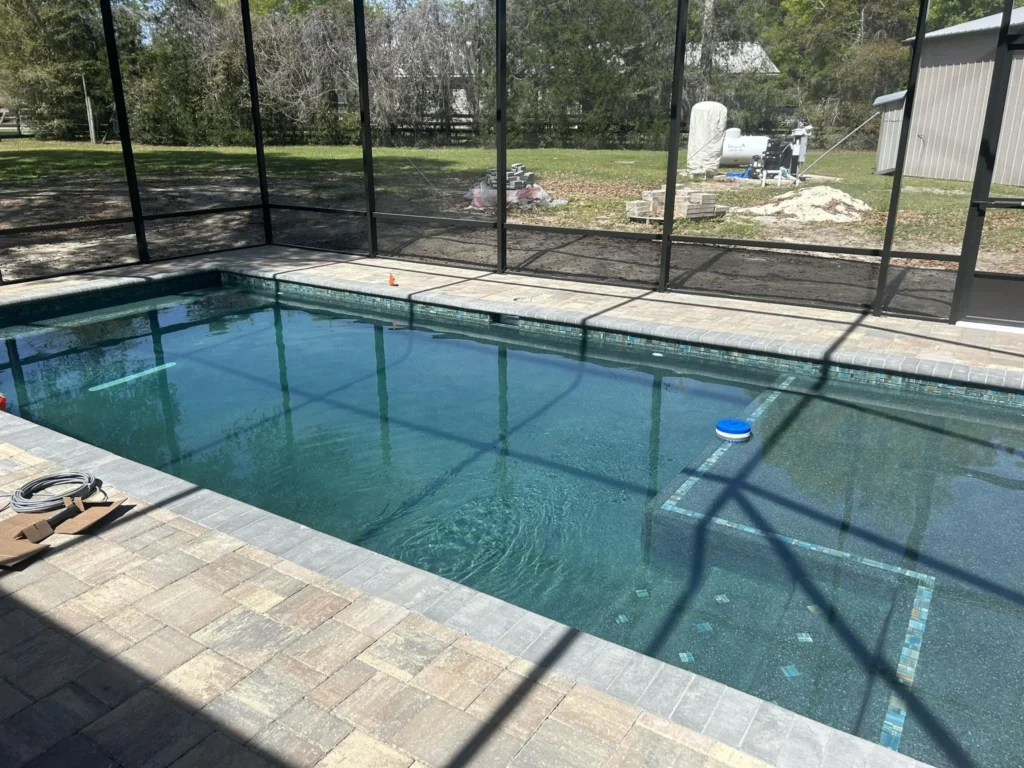 A newly built rectangular pool under a screen enclosure with paver decking, backyard still under construction.