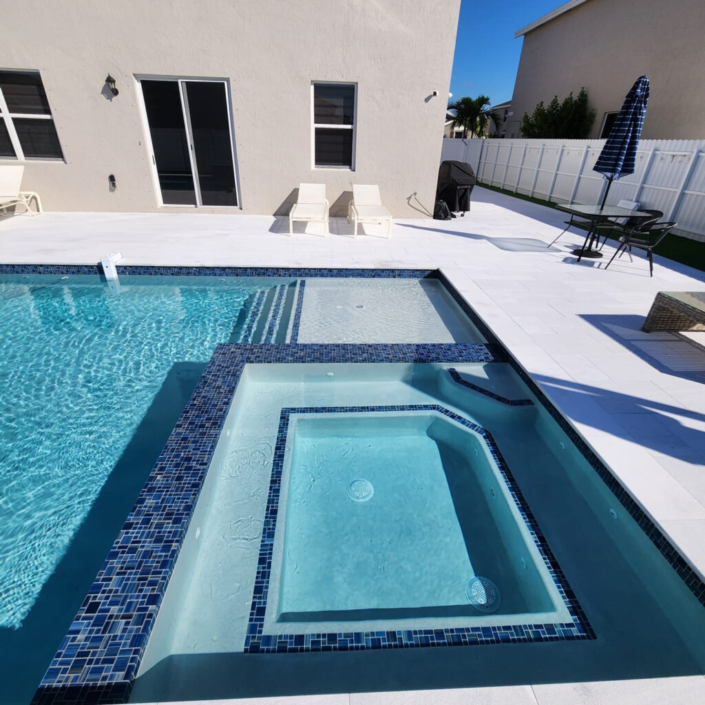A raised spa with blue mosaic tile trim sits beside a pool on a light paver patio.