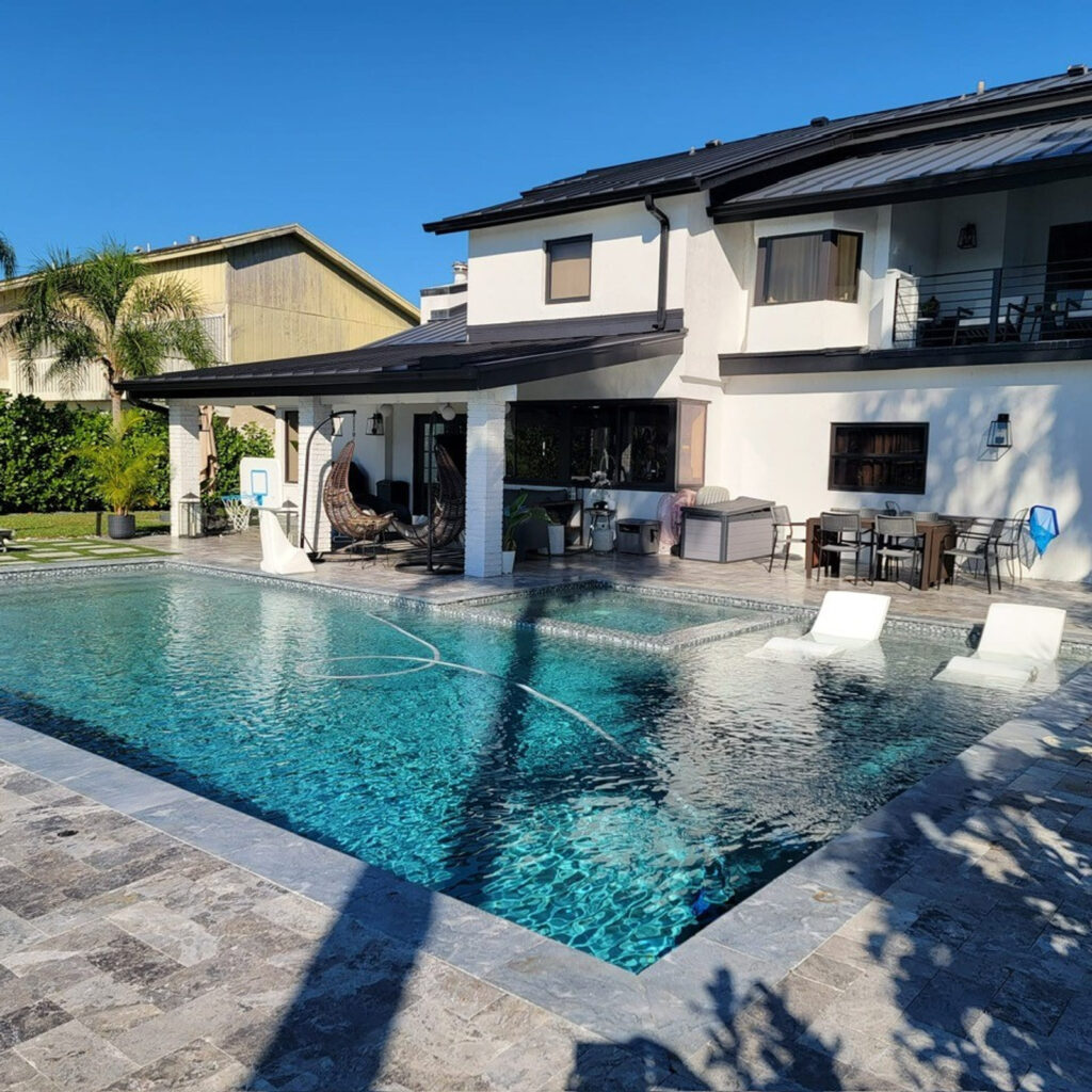 A rectangular backyard pool with an attached sun shelf and two white in-pool loungers in front of a two-story white house.