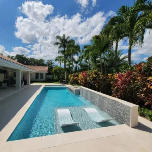 A rectangular backyard pool with two in-water loungers and a raised stone accent wall with jets, surrounded by tropical landscaping.