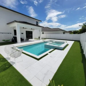 A rectangular backyard pool with an attached raised spa, white pavers, and artificial turf under a bright blue sky.