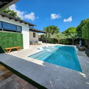 A small rectangular backyard pool with a submerged tanning ledge and loungers, bordered by travertine decking, palm trees, and a decorative water bowl fountain.