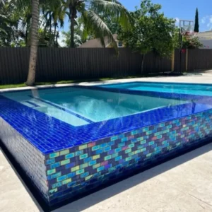 An elevated rectangular pool with a striking iridescent glass tile exterior wall in a fenced backyard with palm trees.