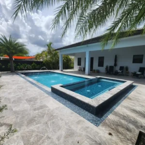 A rectangular pool with an attached spa edged in dark mosaic tile, set on a travertine paver patio behind a house.