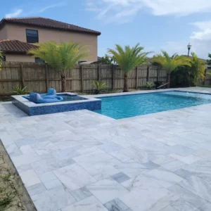 A rectangular pool with a raised spa and blue glass tile, surrounded by a travertine deck, palm trees, and a wood privacy fence.