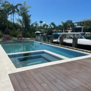 A rectangular pool with an attached raised spa on a canal-front deck with boats docked nearby.
