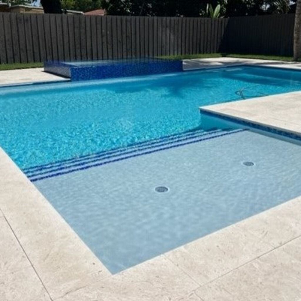 An L-shaped in-ground pool with a raised tiled water feature wall and a shallow sun shelf in a fenced backyard.