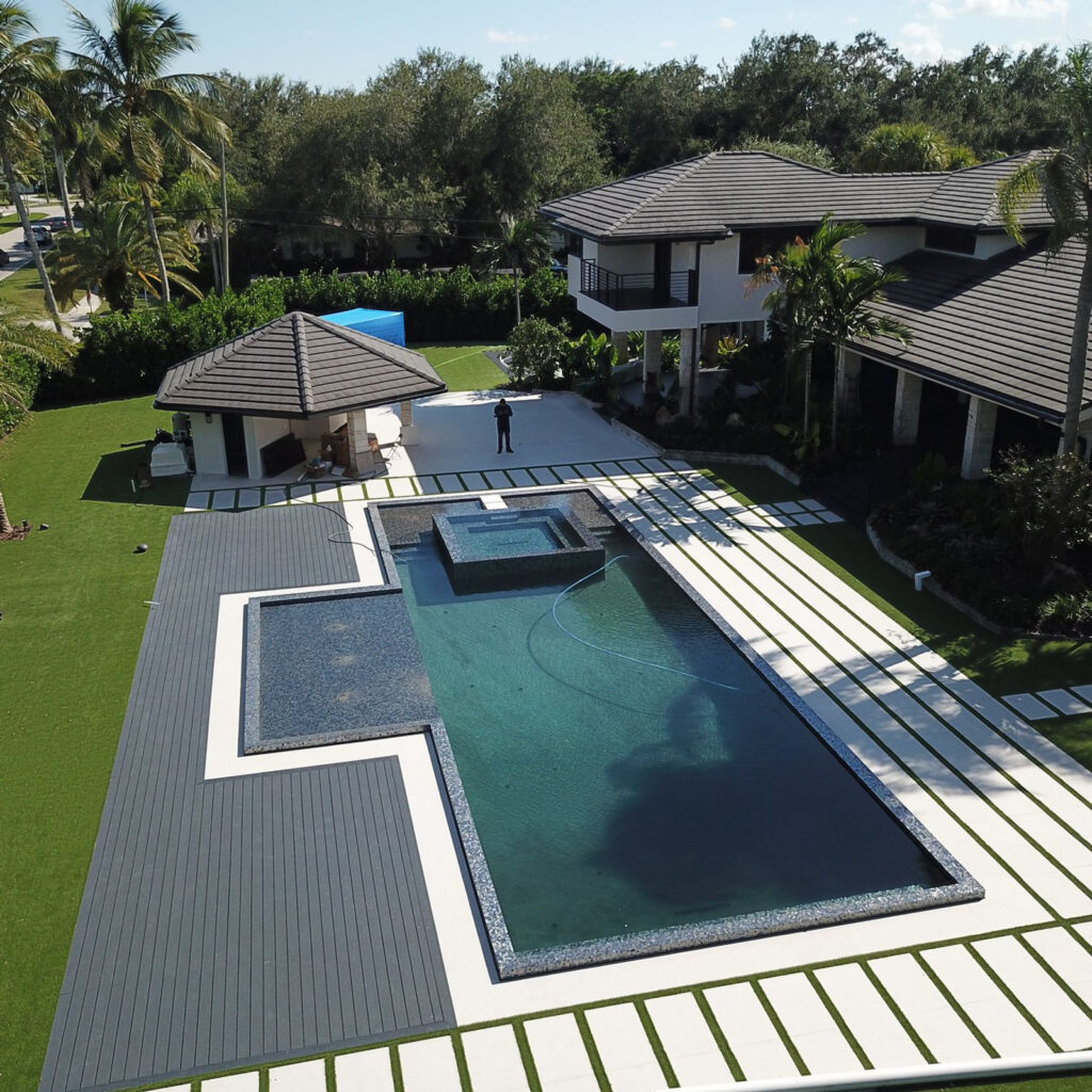 Aerial view of a large modern rectangular pool with an attached raised spa and tanning ledge beside a luxury home.