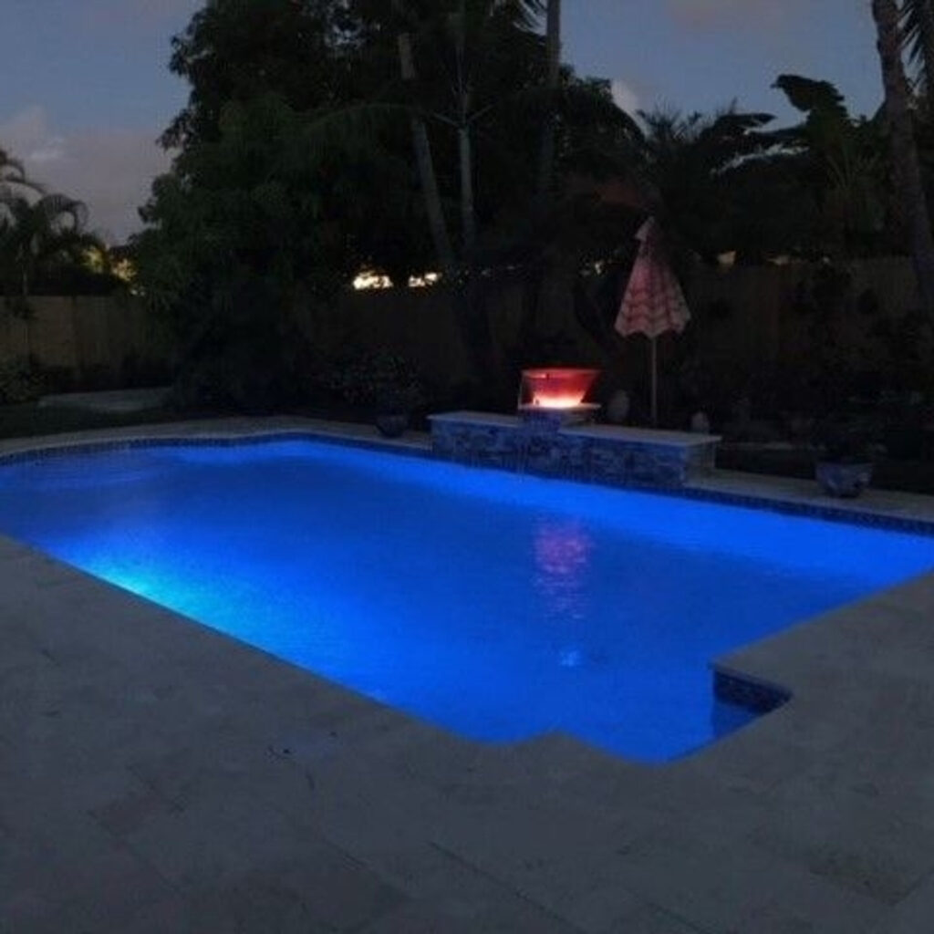 A rectangular backyard pool glowing blue at night with a lit red water bowl feature on a raised wall.