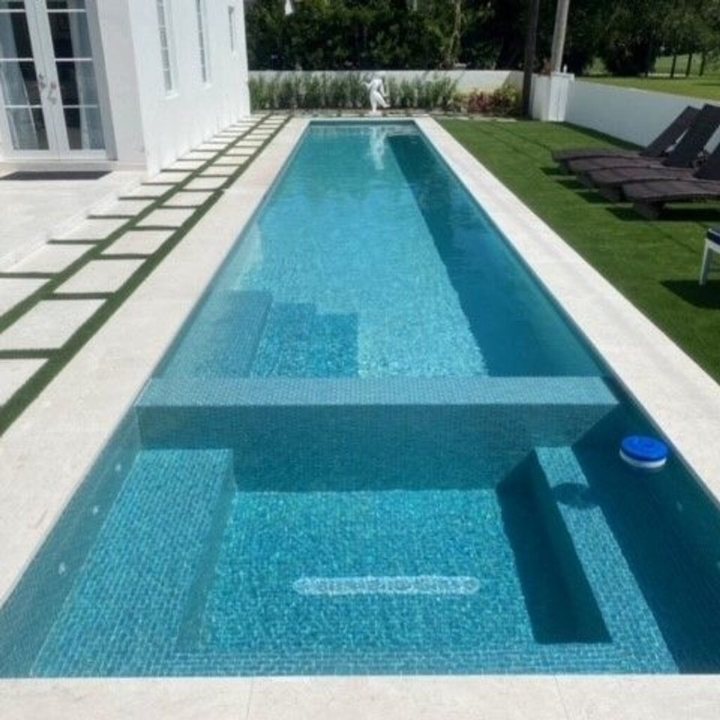 A long narrow lap pool with an all-tile turquoise interior runs alongside a modern white house.