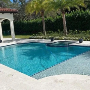 A rectangular pool with a raised spa spilling into it, surrounded by travertine decking and palm trees.