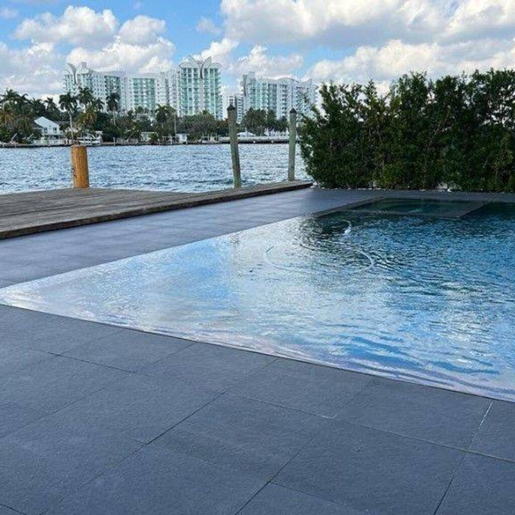 A modern geometric pool corner with a flush spa, overlooking a waterway with a dock and city skyline.