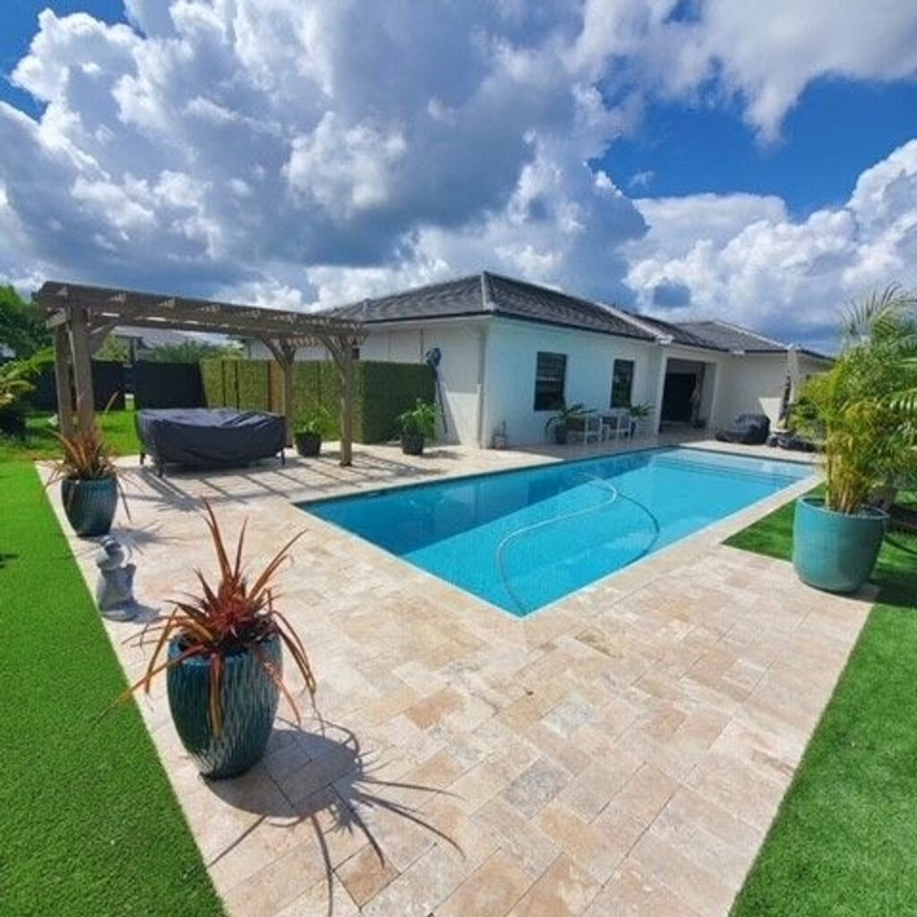 A rectangular backyard pool with a travertine paver deck, pergola, and artificial turf under a partly cloudy sky.