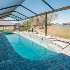 A freeform pool with a tiled raised wall and fountain jet under a screened lanai overlooking a golf course pond.