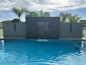 Rectangular backyard pool with a modern raised wall waterfall feature framed by palm trees.