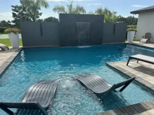 A rectangular backyard pool with a raised water wall feature and two submerged in-pool loungers on a sun shelf.