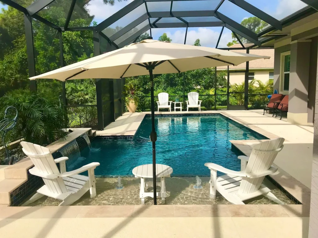 A screened lanai with a small freeform pool featuring a sheer descent waterfall and Adirondack chairs on a travertine patio.