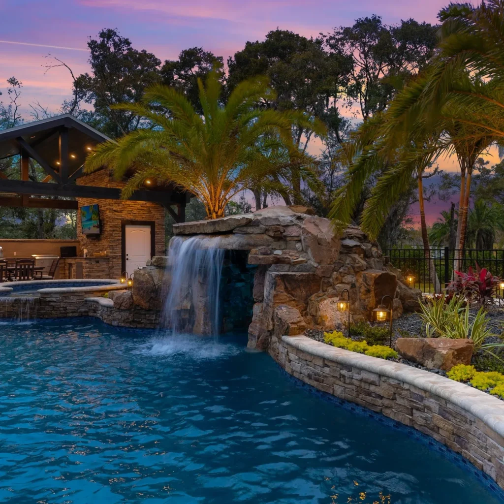 A dusk view of a pool with a stone waterfall grotto, raised spa, and covered outdoor kitchen surrounded by palm trees.