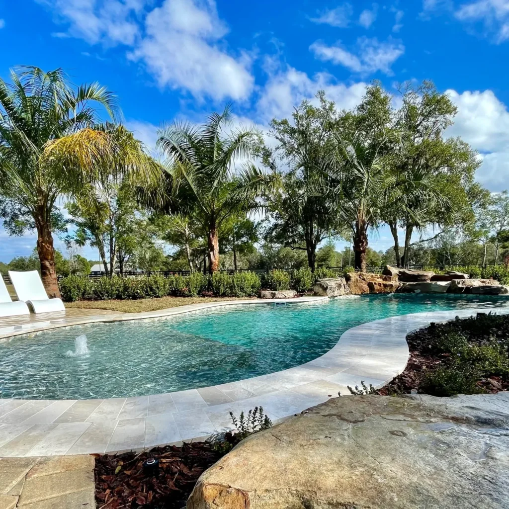 A freeform pool with a bubbler jet and natural rock waterfall surrounded by palm trees and travertine decking.