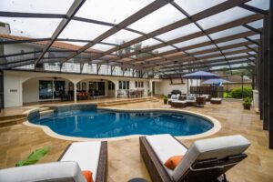 A freeform pool with a stone deck under a large screened lanai behind a two-story house.