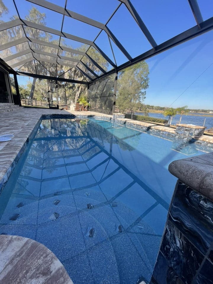 A screened lakefront pool with a long rectangular lap area and dark tiled raised spa edge.